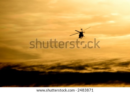 A helicopter flying above a layer of dramatic clouds.  It is late in the day. There is a warm glow to the clouds and sky.  The helicopter is lit from underneath with reflected warm sunlight.