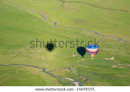 Horizontal photo with shadow of two hot air balloons and their baskets cast on ground below and actual balloon on right