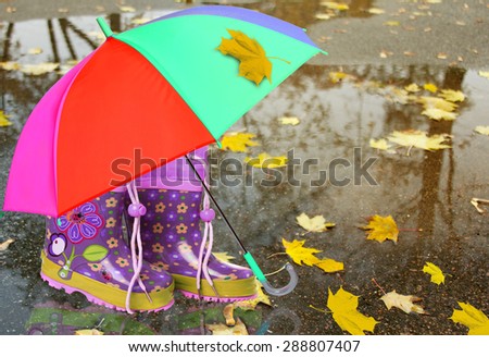 Pink rubber boots with floral print and a colorful umbrella outdoors in rainy weather. Set for a child, for the girls for a comfortable walk in the rain. Symbol of protection, custody.