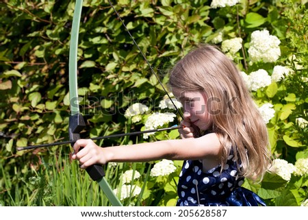 little caucasian girl aiming arrow from big bow on green garden outdoor background