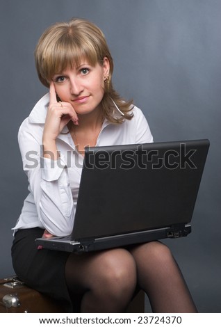 young thoughtful business woman in white shirt and black skirt sitting on suitcase with notebook on her knees on grey background