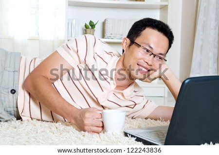 Asian man lying on the floor holding a cup of drink and surfing internet