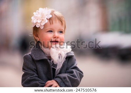 portrait of little baby girl in gray coat on the street in old city with funny smile. One year old girl dressed on grey coat with big white flower in her hear and big white bow around neck.
