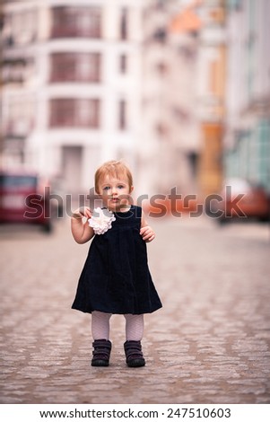 little lady stand on the street. One year old girl dressed on dark velvet dress with big white flower and white tights. The background is diffused with yellow and brown colors