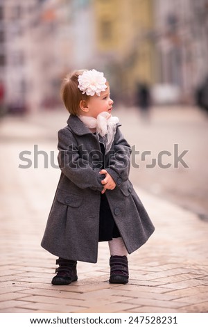 portrait of little baby girl in gray coat on the street in old city with funny smile. One year old girl dressed on grey coat with big white flower in her hear and big white bow around neck. look back
