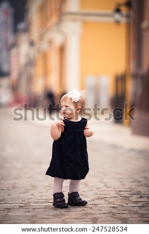 little lady stand on the street. One year old girl dressed on dark velvet dress and white tights with big white flower in hear. She looks back. The background is diffused with yellow and brown colors