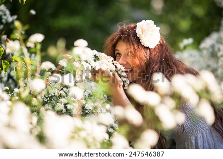 Portrait of beautiful young lady in spring garden full of white flowers. She touches branches and flowers by hands. Young woman in white dress with curly hear and big white flower in her hear