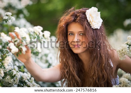 Beautiful brunette lady in spring garden full of white flowers touches branches and flowers by hands.Young woman in white dress with long curly hear and big white flower and petals in hear