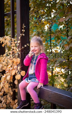 Smiling funny little girl in bright clothes picking her nose. She sits on the brown wooden fence. Bright orange leaves around her.