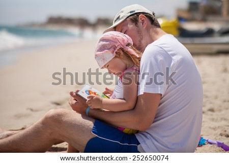 Father and daughter on the beach near the sea. Young man hugs little baby girl and reads her a book