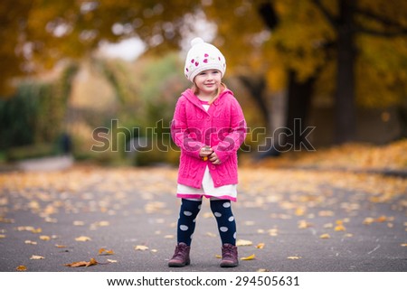 Little girl in bright clothes stand  in autumn garden. Bright autumn alley with yellow leaves on background. Baby puts her hands together and smiles