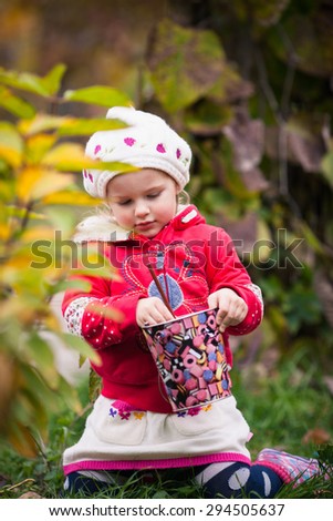 Little girl in bright clothes sits on the grass in autumn garden.  Baby holds colored pencil and bright colored pail