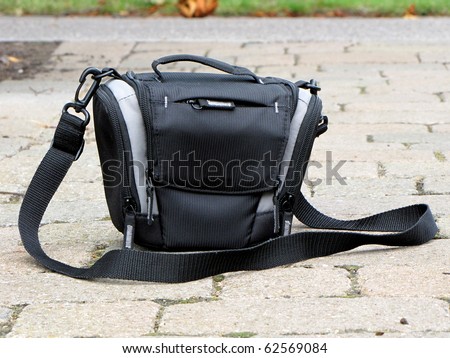 Black camera bag, zipped, standing on cobblestone pavement.