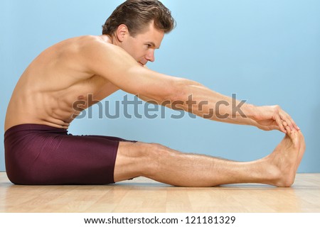Athletic shirtless man performs sitting hamstring stretch by touching toes on wood floor