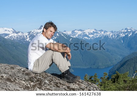 Caucasian man sits on rock on mountain peak overlooking beautiful southeast Alaska landscape including Silver Bay and mountains of Baranof Island on clear summer day