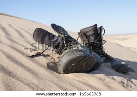Old used black military boots lie half buried in desert sand dune under clear sunny sky