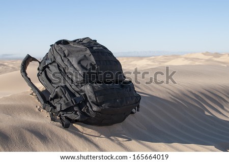 Loaded black military backpack sits on top of desert sand dune under clear sunny sky