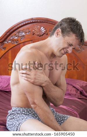 Muscular Caucasian man painfully clutches shoulder with hand as he sits in bed