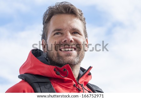 Closeup of happy expression of bearded male Caucasian hiker outside in red jacket smiling with partly cloudy sky background