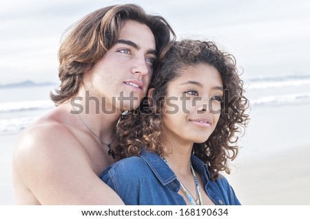 Young Caucasian man embraces young pretty Hispanic woman as they smile while gazing on beautiful beach