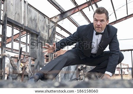 Handsome tough Caucasian man in black tuxedo poses in action stunt scene in destroyed warehouse