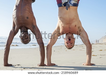 Muscular lean black man and white man perform handstand together on sandy beach under clear sunny sky