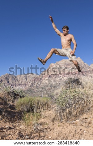 Muscular shirtless Caucasian man wearing shorts and sandals runs and jumps high in air over bushes in harsh desert landscape under clear sunny blue sky in Red Rock Canyon, Las Vegas