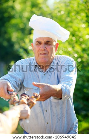 Mature Caucasian man in chef\'s hat holding grilled shish kebabs on skewers, outdoors