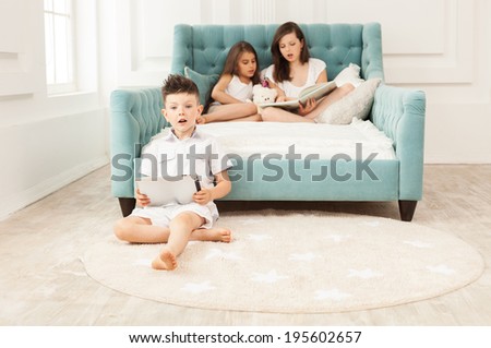 Siblings spending free time at home: two girls reading book on couch and boy using tablet while sitting on floor