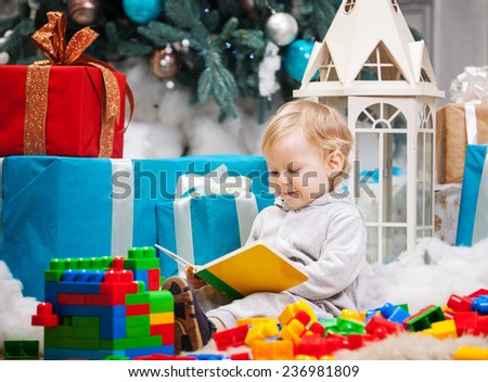Cute toddler boy sitting at Christmas tree and reading book. Building blocks scattered around.