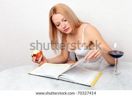 Beautiful young woman eating apple while reading book, with glass of red wine on the table