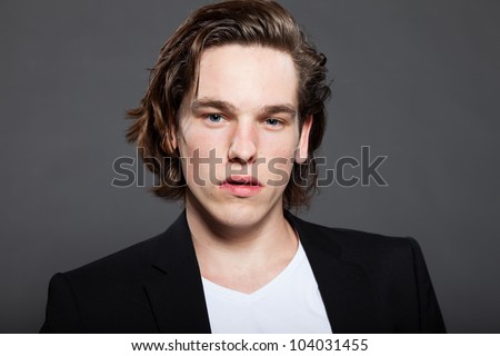 Handsome young man with brown long hair and wearing white shirt and blue jacket isolated on grey background. Fashion studio shot. Expressive face.