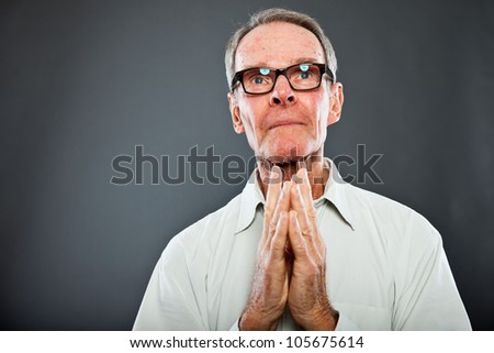 Expressive good looking senior man with glasses against grey wall. Hands praying. Spiritual and characteristic. Well dressed. Studio shot.