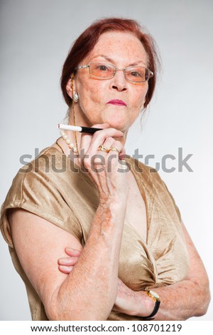 Portrait of good looking senior woman wearing glasses with expressive face showing emotions. Smoking a cigarette. Acting young. Studio shot isolated on grey background.