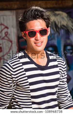 Urban asian man with red sunglasses. Good looking. Cool guy. Wearing blue white striped sweater. Standing in front of wooden wall with graffiti.