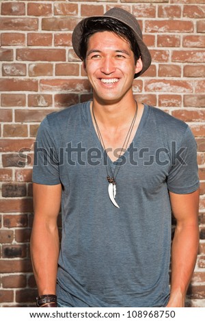 Urban asian man. Good looking. Cool guy. Wearing grey shirt and hat. Standing in front of brick wall.