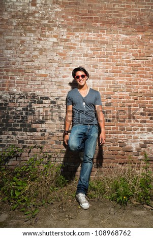 Urban asian man with red sunglasses. Good looking. Cool guy. Wearing grey shirt and hat and jeans. Standing in front of brick wall.