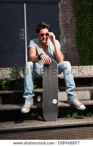 Urban asian man with red sunglasses and skateboard sitting on stairs. Good looking. Cool guy. Wearing grey shirt and jeans. Old neglected building in the background.