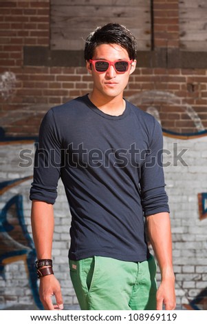 Urban asian man with red sunglasses. Good looking. Cool guy. Wearing dark blue shirt and green shorts. Standing in front of brick wall with graffiti.
