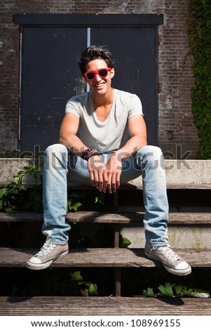 Urban asian man with red sunglasses sitting on stairs. Good looking. Cool guy. Wearing grey shirt and jeans. Old neglected building in the background.