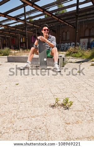Urban asian man with red sunglasses and skateboard sitting on street. Good looking. Cool guy. Wearing blue white striped sweater and green shorts. Old neglected building in the background.