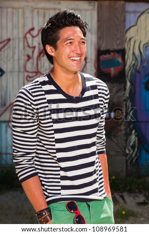 Urban asian man. Good looking. Cool guy. Wearing blue white striped sweater and green shorts. Standing in front of wooden wall with graffiti.