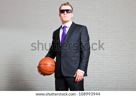 Business man with basketball. Wearing dark sunglasses. Good looking young man with short blond hair. White brick wall.