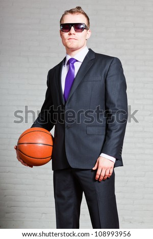 Business man with basketball. Wearing dark sunglasses. Good looking young man with short blond hair. White brick wall.