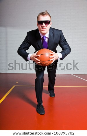 Angry business man with basketball. Wearing dark sunglasses. Good looking young man with short blond hair. Gym indoor.