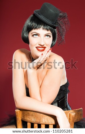 Burlesque pin up woman with black hair dressed in black. Sexy pose. Sitting on chair. Wearing black hat. Studio fashion shot isolated on red background.