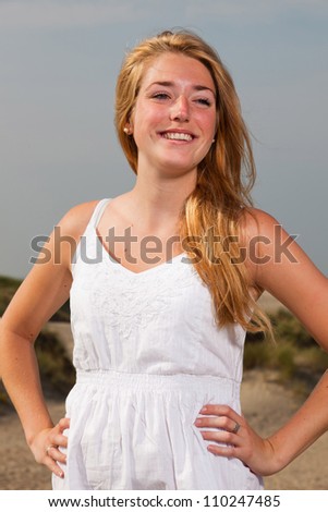 Pretty girl with red long hair wearing white dress enjoying nature near the beach. Hot summer day with blue cloudy sky.