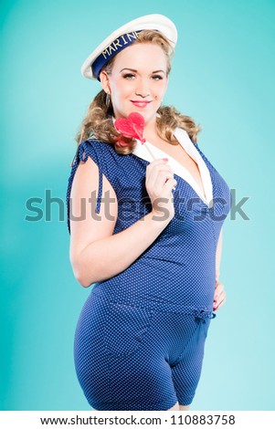 Sexy blonde pin up girl wearing blue dress with white dots and marine cap. Holding pink lollypop. Retro style. Fashion studio shot isolated on light blue background.