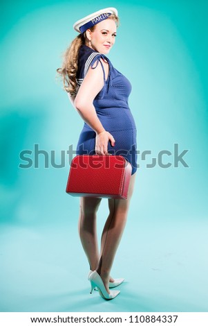 Sexy blonde pin up girl wearing blue dress with white dots and marine cap. Holding vintage red suitcase. Retro style. Fashion studio shot isolated on light blue background.