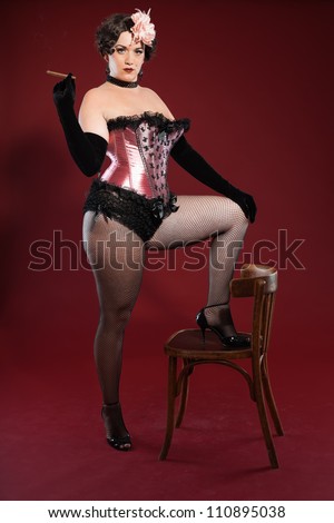 Sexy burlesque pin up woman with long blond hair dressed in pink and black. Smoking cigar. Standing on retro wooden chair. Studio fashion shot isolated on red background.
